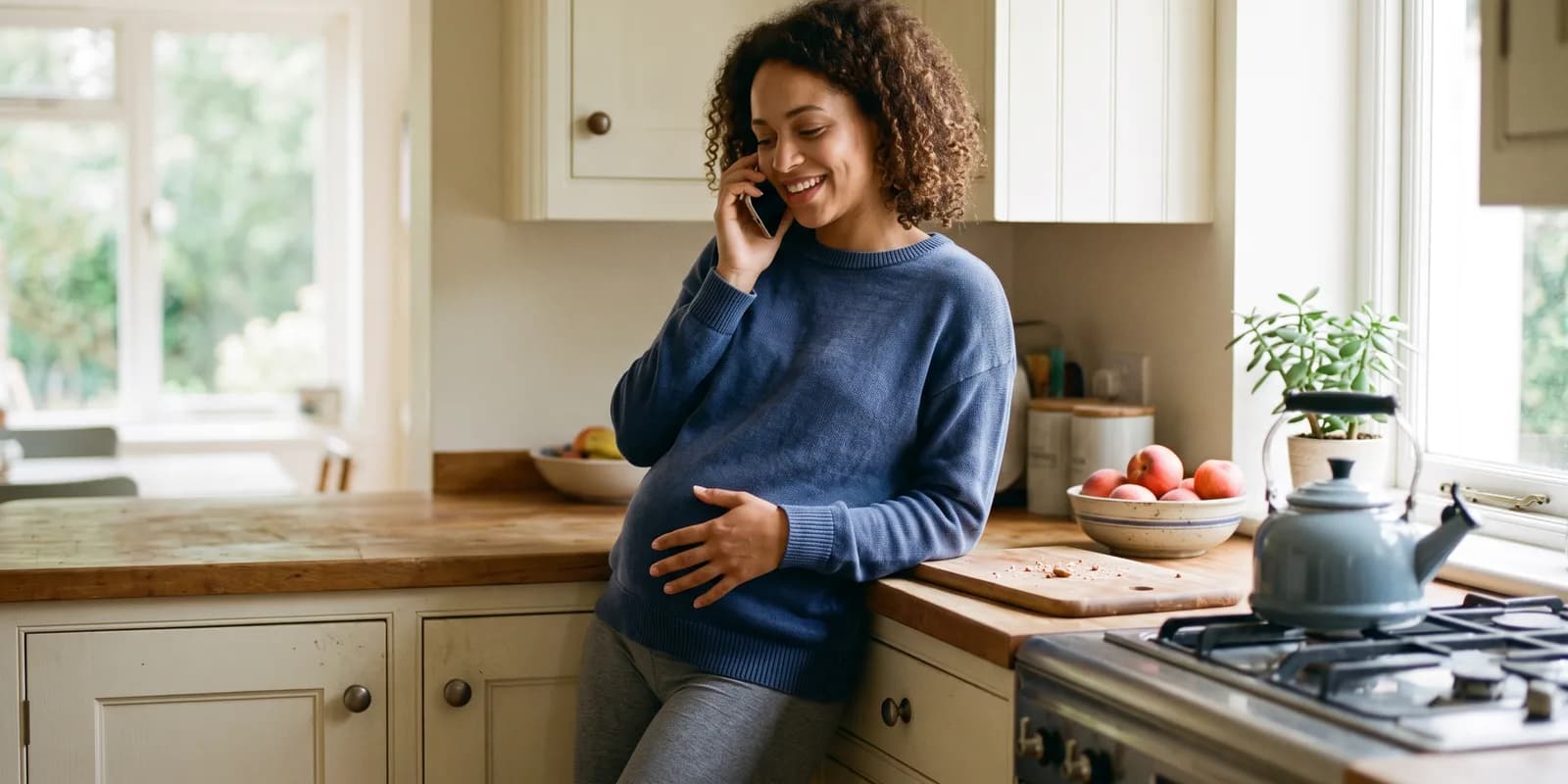 Calling an adoption counselor about a second pregnancy A pregnant woman leans against her kitchen counter on a phone call, one hand on her belly, smiling in morning light